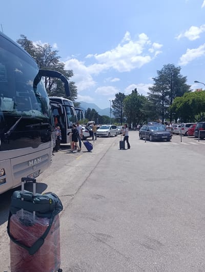 In Mrčevac, Montenegro, on a sunny day, a parked bus, identified by the "MAN" logo and license plate "TVO AZ 849," is in the foreground. Next to it, another bus is parked with passengers around it. A group of people are gathered near the second bus, with some holding luggage. One person is seen pulling a blue suitcase. In the background, several cars are parked, including a dark grey Volkswagen sedan with license plate "TV-TX054". Beyond the parking lot, trees line the area, and mountains are visible in the distance under a clear blue sky with scattered clouds. A streetlight is also present in the background.