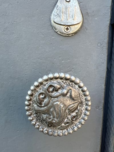 The image shows a close-up of a gray, painted door with two ornate metal elements. The top element is a silver-toned metal fixture with vertical grooves and a screw holding it in place. Below it, a round, silver-toned metal knob features intricate detailing, possibly depicting a creature or mythological figure. The knob is surrounded by a beaded border. To the right of the door, a dark, textured surface, possibly a fence or grate, is visible. The scene is devoid of human presence and activity. The image was taken outdoors, as suggested by the lighting and textures. No specific landmarks or architectural styles are identifiable that would pinpoint a location in Tashelka, Russia.