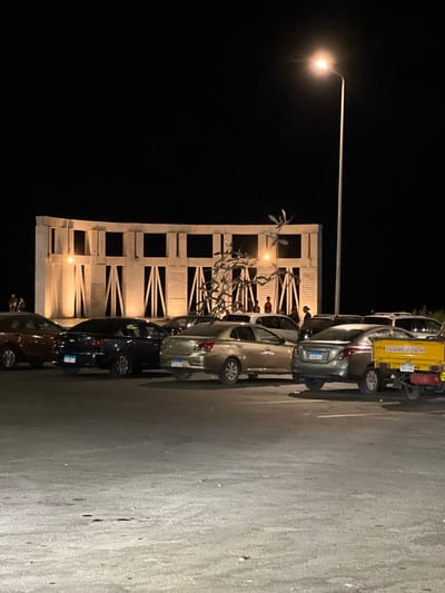 The image depicts a nighttime scene in Sharm El Sheikh, Egypt, featuring a street lamp illuminating a parking area adjacent to a modern, white monument. Several vehicles are parked in the area, including a yellow three-wheeled truck and multiple cars. The monument has a distinct architectural style with multiple rectangular sections and decorative metalwork in the foreground. People are visible near the monument, suggesting an event or gathering. The lighting from the street lamp casts long shadows and highlights the textured surface of the asphalt parking lot. The overall atmosphere is dimly lit, characteristic of nighttime in a populated area. No specific weather conditions are apparent, but the clarity of the image suggests clear weather. The visible text on the license plates of the vehicles is in Arabic script, reinforcing the location context.