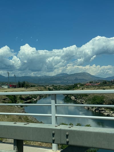 The image depicts a river flowing through a landscape in Podgorica, Montenegro. A bridge railing, constructed from white metal bars and a grey metal beam with visible bolts, dominates the foreground. The river, reflecting the sky, flows through a rocky, vegetated gorge. In the middle ground, dry grass covers rolling hills. Further back, on the left, are two tall stadium lights and a red building. A cluster of cypress trees stands between the lights and the background. In the distance, a range of mountains under a cloudy blue sky forms the horizon. Several buildings with red roofs, some with solar panels, are visible on the hills in the middle to far ground. The scene appears to be a sunny day.