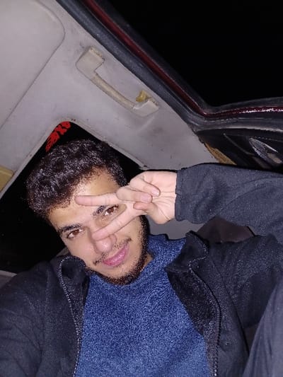 An individual is seen from a slightly elevated angle, possibly inside a car. He is making a hand gesture resembling a peace sign in front of his eye. He wears a blue textured sweater and a dark jacket. The interior of what appears to be a car is visible, including a handle and part of the roof. The background shows a dark space with minimal light.