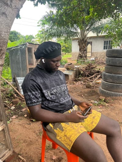 An adult male sits in an orange plastic chair in an outdoor location, appearing to be in Markurdi, Nigeria. He is wearing a black patterned shirt, yellow patterned shorts, and a black head covering. He has an earbud in his left ear and is holding a smartphone in his hands, apparently focused on it. Behind him are stacked tires, a palm tree, and a building. The ground is dirt with scattered vegetation.