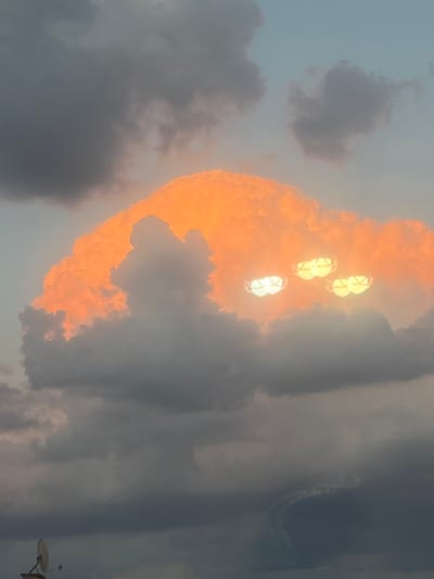In the evening sky above Cheshnegirovo, Bulgaria, a large, luminous cloud formation is visible, tinged with vibrant orange from the setting sun. Embedded within this cloud are three distinct, glowing orbs that resemble spotlights, casting a bright, diffused light. These orbs are positioned within the cloud, appearing almost as if they are part of its structure or illuminating it from within. Below this prominent formation, darker, more diffused clouds fill the lower portion of the frame. In the bottom left corner of the image, a satellite dish mounted on a structure is partially visible, suggesting a human presence or habitation in the area. The overall scene captures a dramatic interplay of light and cloud during twilight.