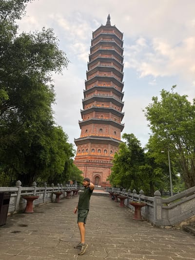 A man wearing a dark green t-shirt and shorts stands in the foreground of an image that features a tall, multi-tiered pagoda in Tây Hoa Lư Ward, Vietnam. The pagoda, constructed of reddish-brown brick with gray roofing, dominates the background. It is adorned with decorative elements and has numerous windows, some illuminated. The man is facing to the right, with his left hand on the back of his head and his right arm slightly bent. He is standing on a paved walkway made of stone slabs.
The walkway is flanked on the left by a stone railing with decorative carvings and several urn-like planters. On the right, a set of stone stairs leads upward. Lush green trees with dense foliage are visible on both sides of the walkway and behind the pagoda. The sky above is overcast with scattered clouds. The overall scene suggests a historical or religious site within a natural setting.