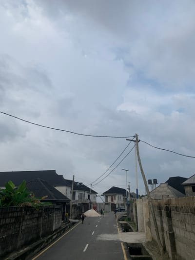 A street scene in Iwofe, Nigeria, is depicted on an overcast day. The upper half of the image is dominated by a cloudy sky, filled with thick grey and white clouds. Electrical wires stretch across the sky, connected to a prominent utility pole positioned towards the center-right of the frame.

An asphalt road runs through the lower portion of the image, extending into the background. It features white dashed lines for lane markings and a yellow line along its right edge. Multi-story buildings line both sides of the street. On the right, structures with dark roofs and concrete walls are visible, one of which has two black water tanks on its rooftop. On the left side, more buildings are present, some appearing finished while others show exposed blockwork, hinting at construction or renovation.

Several individuals are on the street. A few people are gathered near a large white pile of material, possibly sand or gravel, on the left side of the road. Further down the street, indistinct figures and a dark-colored car can be seen. Green foliage is visible in the bottom right foreground. The scene portrays an active urban or semi-urban environment under a uniformly cloudy sky, with no direct sunlight. No discernible text is visible within the image.