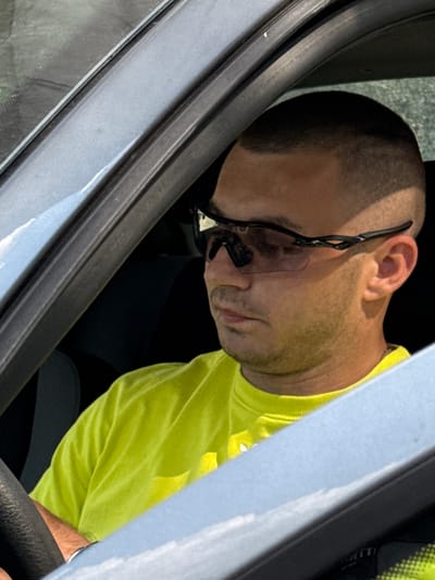 The media file is a close-up, candid shot of a person in a vehicle, likely a truck. The subject is a man with a buzzed haircut, wearing safety glasses pushed up onto his forehead and a bright yellow t-shirt. His head is tilted downwards, and his eyes appear closed, suggesting he might be sleeping or resting. The man is seated in what appears to be the driver's seat, with a black steering wheel partially visible in the foreground.

The scene is framed by the car's interior, with the dark rubber seal of the door and window visible on the left and the light-colored interior panel on the right. The lighting suggests it is daytime, with natural light coming from outside the vehicle. The overall mood is one of rest or inactivity. There are no visible landmarks or text that provide specific location cues beyond being inside a vehicle. The provided location context is Shumen, Bulgaria, which could be the general area where the photo was taken.