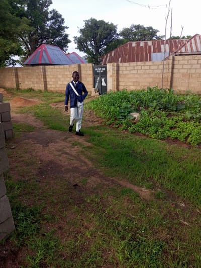 A young man dressed in a dark blue jacket, white shirt, and white pants with a white sash walks on a dirt path in Zinna, Nigeria. He is wearing black shoes. To his right is a garden with green plants. Behind the garden is a brick wall. Beyond the wall are two structures with red and grey corrugated metal roofs, and a building with a rusted corrugated metal roof. A sign on a green gate reads "MILITARY ZONE KEEP OFF" and depicts a dog. The sky is overcast.