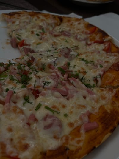 A close-up, high-angle shot captures a partially consumed pizza resting on a white, round plate in what is identified as the city of Novopokrovskaya, Russia. The pizza features a golden-brown crust and is generously covered with melted white cheese, displaying slight browning in various spots. Distributed across the cheese are numerous pieces of light pink, processed meat, consistent with ham or sausage, cut into irregular strips and chunks. Red bell pepper segments, varying in size, are also visible among the other toppings. Fresh green herbs, primarily dill and possibly parsley, are sparsely sprinkled over the entire surface. One slice of the pizza has been removed from the left side, leaving an empty space on the plate. The background is blurred, showing faint outlines of other objects, suggesting a dining setting.