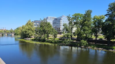 The image depicts a serene waterside scene in Linköping, Sweden, likely near the Kinda Canal or the river Stångån, given the urban setting and the presence of modern apartment buildings. The foreground is dominated by a calm body of water, its surface reflecting the clear blue sky and the surrounding greenery. Lily pads are scattered across the water, indicating a shallow or calm area.
Along the bank, a paved walkway runs parallel to the water, bordered by lush green grass and mature trees. The trees, with their full, vibrant foliage, create a natural canopy and provide shade. Behind the trees, a series of modern, multi-story apartment buildings are visible. These buildings feature contemporary architectural styles with large windows and balconies, suggesting a well-developed urban residential area. One building has a white, patterned facade on its upper floors, adding a unique visual element.
In the mid-ground, a small wooden jetty extends into the water, and a single person can be seen standing on it, possibly preparing to swim or simply enjoying the view. Further in the distance, a bridge crosses the waterway, and beyond that, industrial-looking chimneys are visible, hinting at the city's historical industrial past, juxtaposed with its modern development. The overall atmosphere is one of peaceful urban coexistence, with nature and modern architecture harmoniously integrated. The scene is bathed in bright sunlight, indicative of a clear, warm day.
