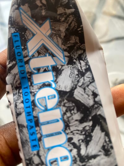 A hand is holding a tube of "Xtreme Fluoride Toothpaste." The tube has a black and white, abstract, rocky pattern as its background. The brand name "Xtreme" is in large, white, capitalized letters with a blue outline. Below the brand name, on a blue stripe, the words "Fluoride Toothpaste" are written in white, capitalized letters. The image appears to be taken indoors, possibly in a home or a shop, given the lighting. The hand holding the toothpaste suggests it is being presented or examined. The overall scene is a close-up of the product, emphasizing the branding and product type. The location context of Jauro Yinu, Nigeria, is provided but not visually evident in the image itself.