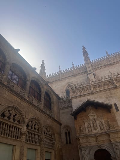 The image shows an exterior view of the Cathedral of Granada, Spain, on a clear day with the sun partially obscuring the top left of the frame. The architecture is Gothic and Renaissance, with intricate stone carvings, arched windows, and decorative pinnacles. The building is made of light-colored stone. In the foreground, a portion of a balcony with balusters and decorative carvings is visible. Above and to the right, the intricate facade of the cathedral rises, featuring multiple tiers of decorative elements and Gothic spires. A small, dark-tiled roof juts out from the facade in the mid-right section of the image, above an ornate portal with religious sculptures. The sky is a pale blue, with a few small birds visible in the upper portion. The sun's glare creates a bright, hazy effect on the left side of the image.