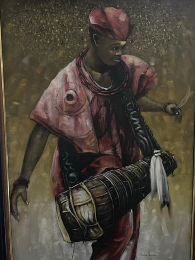 This image is a photograph of a framed oil painting depicting a single individual, likely male, positioned slightly off-center and angled to the right. The subject wears a red traditional cap (fila) and a loose, reddish-pink tunic adorned with circular patterns and darker intricate designs. A dark, patterned strap crosses the subject's chest, securing a brown elongated talking drum (gangan) that rests across the lower body. The drum features white wrappings and decorative tassels. The individual holds two thin, curved drumsticks in the raised right hand and wears red wrist bracelets on both arms. The left hand is slightly open and lowered. The subject's dark skin tone is visible, and the gaze is directed towards the right. The background is an abstract, textured field of shimmering gold, brown, and ochre tones with speckled highlights. The painting is enclosed within a dark frame featuring a thin gold inner border. A signature, partially visible as "Chuks Okonkwo" or similar, is located in the bottom right corner of the canvas. This painting is located in Ikate, Nigeria.
