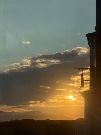 The image captures a sunset or sunrise over a mountainous landscape, with the sun appearing as two bright orbs partially obscured by clouds. The sky transitions from a clear, light blue on the left to a warm, golden hue on the right, illuminated by the setting or rising sun. Wispy clouds are scattered across the sky, catching the sunlight and creating dramatic contrasts between light and shadow.
In the foreground, the silhouette of a building or structure is visible, with dark edges and some indistinct details. A horizontal bar with strings attached, possibly a clothesline, is also silhouetted against the bright sky. In the distance, rolling hills or mountains are visible, also appearing in silhouette against the vibrant sky. The overall scene evokes a sense of tranquility and the beauty of nature's display. The time of day is clearly indicated by the golden light and the position of the sun. The weather appears clear with some cloud cover. There is no visible text in the image. The location context is Petropavlovsk-Kamchatsky, Russia.