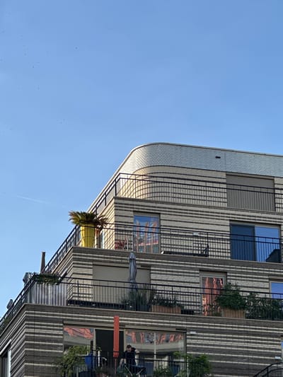 This is a vertical, eye-level medium shot of a modern apartment building's facade on a clear, sunny day. The building, likely located in Amsterdam, Netherlands, has a distinctive patterned facade composed of light-colored bricks with dark vertical accents. The building curves slightly on the left side of the frame, creating an interesting architectural detail.
The image captures several balconies with black metal railings. One prominent balcony features a large yellow planter with a bushy green plant. Other balconies show glimpses of urban life: plants, outdoor furniture, and what appears to be an umbrella. Reflections of a warm, possibly sunset, sky are visible in the building's windows, suggesting the photograph was taken in the late afternoon or early evening. A thin contrail streaks across the clear blue sky.
While no people are clearly depicted, one figure can be seen on a balcony in the upper right section of the image, looking away from the camera. The overall scene is one of a peaceful urban environment, bathed in warm sunlight. There is no visible text in the image.