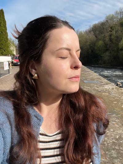 A woman with light skin and long, reddish-brown hair lies on a concrete ledge outdoors. Her eyes are closed, and her face is turned towards the sky. She wears a light blue sweater over a striped shirt. A gold earring is visible in her left ear.

Behind her, a river flows past a wooded area. A concrete wall and a street with a red vehicle and greenery are partially visible in the foreground.

The location is likely in Gangloff, Germany, based on the request context.