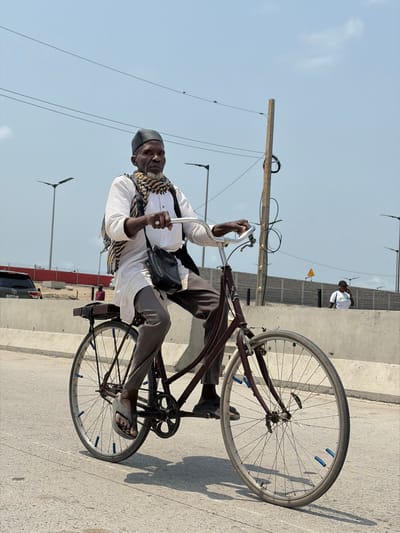 An elderly man of African descent is depicted riding a dark red bicycle on a paved road in Igboefon, Nigeria, under a clear, light blue sky. He wears a black skullcap, a white long-sleeved tunic, grey trousers, and a black-and-white patterned scarf around his neck. A black shoulder bag is slung across his body. He has a ring on the ring finger of his left hand, which grips the silver handlebars. His left foot is in a grey flip-flop, while his right foot wears a dark slip-on shoe. The bicycle features a black seat with a small, dark cargo rack over the rear wheel. Both wheels have silver rims and spokes, with visible blue valve caps on some spokes. Shadows cast on the road indicate direct overhead sunlight. In the background, concrete barriers line both sides of the road. Behind the left barrier, a dark SUV is partially visible, along with red fencing. Behind the right barrier, a person in a white T-shirt and dark cap stands, and a yellow triangular warning sign is affixed to the barrier. Multiple grey streetlights and overhead utility lines with a wooden pole are present in the mid-ground and background.
