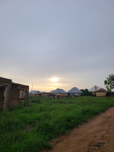 This is a medium-shot, outdoor, vertical photograph taken at dusk in Kuru Karama, Nigeria. The scene depicts a rural or semi-rural area with several houses and a partially constructed building. The sky is overcast with a bright sun partially visible, creating a soft, warm glow.
In the foreground on the left, a building is under construction, with exposed brickwork and concrete pillars. A dirt path runs from the right foreground into the mid-ground.
In the mid-ground, there is a large expanse of tall, green grass. Several houses with metallic roofs are visible, some appearing complete and others still in various stages of development.
In the background, more houses are visible, along with trees and vegetation. The overall impression is one of a developing community in a natural setting. There are no people clearly visible in the frame.