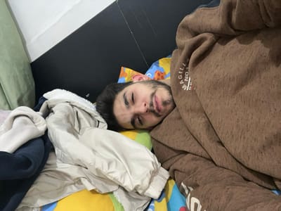 A man lies in bed surrounded by clothes. His face is visible, with eyes partially closed. He is partially covered by a brown hooded sweatshirt. A pile of clothing, including a tan jacket with a white lining and a dark blue garment, sits beside him. The bed has a yellow and blue patterned sheet. The headboard is black. A light green curtain is visible in the upper left corner. The lighting is dim.