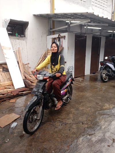 The image shows a woman sitting on a black motorcycle in a grey, concrete driveway. She is wearing a yellow long-sleeved shirt, brown pants, and a black headscarf. She is looking at the camera and smiling. The motorcycle has a black license plate that reads "5541 YAW 05-27." The motorcycle is parked in front of a building with a metal roof. There are two wooden doors to the right of the motorcycle. To the left of the motorcycle is a pile of wood scraps. The scene appears to be in Katapang, Indonesia, as indicated by the provided coordinates. The ground is wet and the weather is overcast.