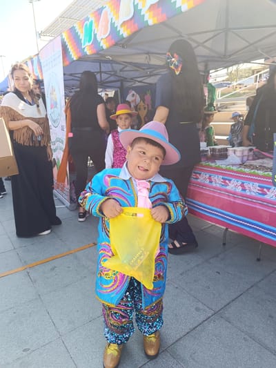 A young boy, dressed in a vibrant blue and gold traditional outfit adorned with sequins, stands smiling and holding a yellow bag. The outfit includes a patterned jacket, pink shirt and tie, and matching sequined pants. He wears a pink and blue hat. He is positioned in the foreground, facing the camera with his mouth open, appearing excited.

In the background, other people are visible, including women and children, some of whom are also dressed in festive attire. Tents and tables are set up, suggesting an outdoor event or market. One tent has a colorful banner with geometric patterns. A table draped with a pink and blue striped cloth displays various items, possibly food or crafts. The ground is paved with grey tiles.

The lighting suggests it is daytime, likely sunny, with clear weather. The overall scene conveys a sense of celebration and community gathering. The setting appears to be a public outdoor space, possibly a fair, festival, or cultural event, potentially in Arica, Chile, given the provided location context.

Visible text includes "CRAYONS" on the yellow bag the boy is holding. There is also text on a banner behind the boy, but it is largely illegible.