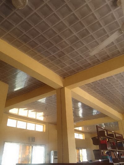 The scene depicts the interior of a building in Nnewi, Nigeria, consistent with a public space such as a library or reading room. The overhead area features a light grey grid-patterned ceiling composed of numerous square recessed panels. A circular light fixture is mounted in the upper-left corner, and a white ceiling fan is partially visible in the upper-right. Thick, light-yellow painted concrete beams and pillars form the primary structural elements, with a prominent intersection occurring in the mid-frame. The walls, also painted light yellow, contain multiple window sets. On the left, a large ground-level window or glass door, fitted with an internal security grill, allows significant natural light into the space. Above this, a row of smaller, rectangular clerestory windows is visible. A white, vertical appliance, likely an air conditioner unit, stands below the larger left window. In the far right background, a dark, multi-tiered shelving unit, consistent with a bookcase, holds various bound materials and storage containers. No human subjects are present. The environment is illuminated by ambient daylight entering through the windows, creating reflections on the ceiling surface.