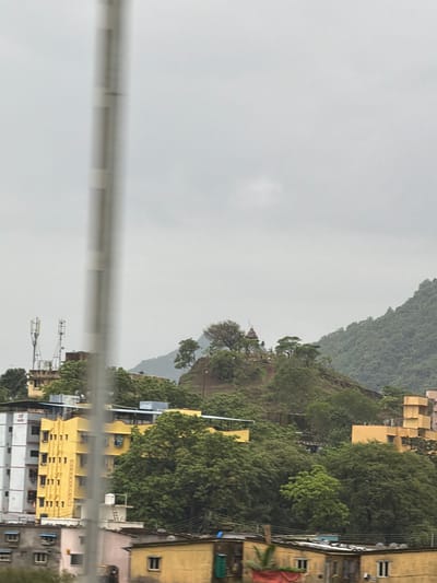 The image depicts a cloudy day in Vasai-Virar, India, with a focus on a residential area nestled against a lush green hillside. In the foreground, several buildings are visible, including a tall, white and gray apartment building with numerous windows and balconies, and a vibrant yellow, multi-story structure with a distinctive architectural design. Adjacent to these are lower-lying, older buildings with various window styles and some visible utilities like satellite dishes.
Further up the hill, more buildings are scattered amongst the dense foliage. A prominent yellow building with a unique, stepped facade stands out. The hillside itself is covered in a thick carpet of trees, with some darker, more rugged areas suggesting steeper inclines or rock formations.
At the crest of a prominent hill, a small, weathered temple or shrine structure is visible, topped with a spire. The surrounding trees on this elevated area are sparser, allowing the structure to be seen more clearly. Two tall communication towers, likely for cellular service, are located on a lower ledge of the hill, to the left of the temple.
The sky is overcast with a uniform layer of gray clouds, obscuring any direct sunlight and casting a diffused light over the entire scene. A thin, vertical, out-of-focus pole-like object runs through the center of the image, partially obscuring the view of the buildings and landscape behind it. The overall impression is one of a densely populated urban environment encroaching on a natural, hilly landscape under a somber sky.
