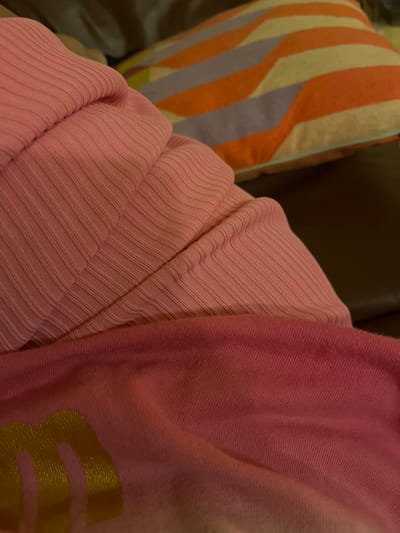 A close-up shot features a pink ribbed sweater overlapping a pink sweatshirt with a gold graphic on the lower right corner. An orange and gray striped pillow is visible on the left, suggesting a domestic setting. The background is dark and blurred, likely part of a sofa or chair. The scene conveys a sense of comfort and relaxation. There is no information to suggest that the image was taken in Santiago de Surco, Peru.