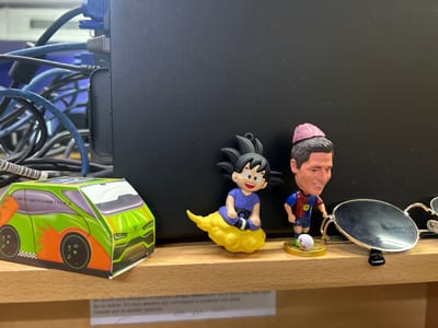 An indoor, eye-level shot captures a collection of decorative and functional items arranged on a light-colored wooden shelf, likely in an office or workspace in Guayaquil, Ecuador. To the left, a colorful cardboard model car, predominantly green with orange accents and black wheels, sits with text "E 2025-10-2027-18 L5-04096 PVP 25" visible on its roof and side.

Central to the image are two small figurines. The first is a young Son Goku from Dragon Ball, wearing a blue gi and sitting on a yellow Flying Nimbus cloud. Next to him is a bobblehead-style figurine resembling footballer Robert Lewandowski, dressed in an FC Barcelona kit (with "Spotify" logo) and a pink hat, standing next to a small soccer ball. To the right of the figurines, a pair of aviator-style sunglasses with dark lenses and a gold-colored frame rests on the shelf.

Behind these items, a black electronic device (possibly a computer tower or server) is visible, with numerous tangled cables (blue, black, and grey, including VGA and Ethernet connectors) plugged into its rear ports. Below the shelf, a piece of white paper is taped to the underside, displaying Spanish text. The legible parts read: "Su tarjeta será entregada en máximo 40 minutos, por favor tenga a mano la copia de su cédula. En unos minutos nos volveremos a contactar con usted. Gracias por su amable atención." No specific activity is occurring, and the time of day is not indicated.
