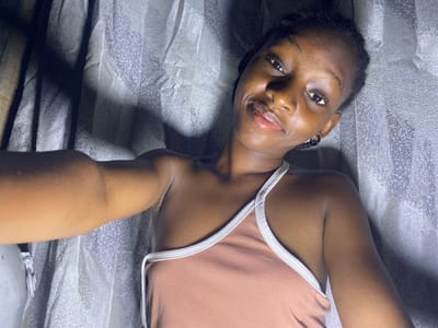 The image shows a single young female, likely an adult, located indoors in Iranla, Nigeria. She has dark skin and her hair is styled in braids. She is wearing a light brown or peach-colored sleeveless top with white trim along the neckline and straps. A black hoop earring is visible in her left ear. The individual is positioned facing the camera, with her head slightly tilted to her left, and she displays a subtle smile. Her right arm is visible in the lower left portion of the frame, extended, suggesting she is holding the device used to capture the image. The background consists of a light gray or silverish textured material, possibly fabric or a patterned wall, with visible shadows indicating an overhead or directional light source.