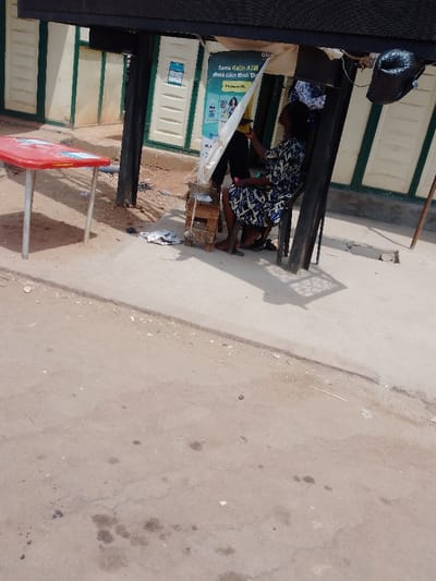 The image displays an outdoor scene in Rido, Nigeria, under clear daylight. The foreground consists of a light-colored concrete or paved surface exhibiting scattered debris, dust, and several dark liquid stains. To the left, a red rectangular plastic table with four silver-colored metal legs stands on this surface.
In the midground, a dark fabric canopy supported by black metal poles covers a small area. Beneath this canopy, a woman is seated on a dark chair or stool, facing slightly towards the right of the frame with her head tilted upwards. She wears a patterned dress, primarily blue, white, and black, along with a matching headwrap. To her left, a makeshift stand or counter constructed from stacked wooden blocks is partially obscured by a white fabric sheet, which extends upwards to hang from the canopy. A portion of another individual's legs and torso, dressed in dark trousers, is visible behind the seated woman. A black, irregularly shaped object hangs from the canopy structure to the right of the seated woman.
The background features building walls painted in a light cream color, accented with green trim or paneling resembling doors. A poster is visible on one of these walls, displaying text including "Samu Katin ATM" and various graphics. Shadows are cast on the ground, indicating the presence of direct sunlight.