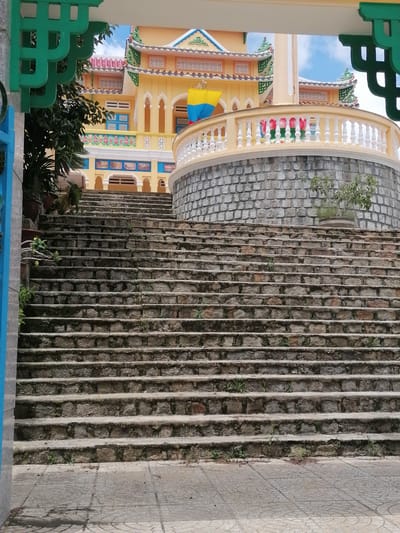 The image shows a stone staircase leading up to a brightly colored temple or large building. The building is yellow with green and blue trim, and features ornate roofs. A white railing with decorative elements lines a curved wall to the right of the stairs. Vegetation is visible growing in pots and along the edges of the stairs. The scene appears to be outdoors, with a blue sky visible above the building. The location is identified as Xuân Thành, Vietnam. There are no people visible in the image.