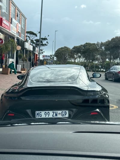 The image shows the rear of a black Aston Martin parked along a street in Cape Town, South Africa. The license plate reads "MG 99 ZW GP".  A building is visible on the left, with signage for "Pizza" and "Hobson's 24/7". Across the street there is an "R+N" sign.  A person wearing a green jacket is visible on the pavement near some cardboard boxes. Another car is parked ahead of the Aston Martin. The sky is overcast and trees line the street. The image was likely taken from inside a vehicle.