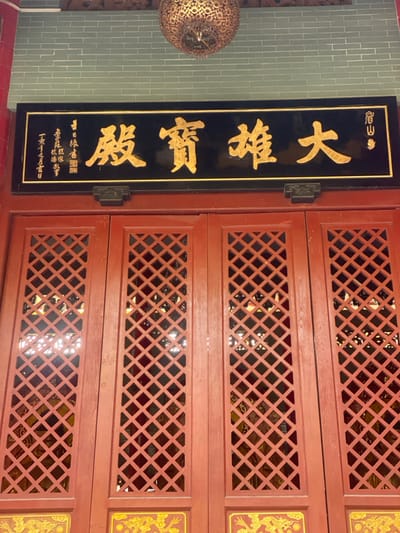 A vertical black plaque with prominent gold Chinese characters dominates the left side of the image, mounted on a reddish-brown wooden structure. The main characters on the plaque, read from top to bottom, are "大慈寶殿" (Dà Cí Bǎo Diàn), translating to "Great Compassion Treasure Hall." Above this, smaller gold characters read "南山" (Nán Shān), meaning "South Mountain." At the bottom of the plaque, even smaller characters indicate a dedication and a date, with the last visible line appearing to be "甲午年九月十八日" (Jiawu year, 18th day of the 9th month). A dark, possibly metal, hinge secures the plaque to the right.

To the right of the plaque are multiple horizontal panels of reddish-brown wood, featuring intricate diamond-shaped lattice patterns. These panels appear to be part of a larger door or wall structure, through which glimpses of an interior space with subtle reflections can be seen. On the far right, partially visible, are sections of vertical rectangular panels with ornate golden patterns, possibly depicting dragons or other auspicious symbols, against a red background.

On the far left, a decorative, spherical object with a complex, possibly metallic or ceramic, golden pattern hangs against a light green or grey tiled wall. No people are visible, and no specific activity is taking place. The scene depicts a static architectural detail, likely within a temple or traditional Chinese building in Guang Zhou Shi, China. The lighting suggests an indoor or sheltered outdoor setting.