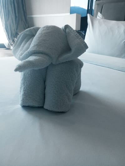 This image depicts a hotel room in Batulawang, Indonesia. A light blue-grey terry cloth towel, meticulously folded to resemble an elephant, is positioned centrally on a neatly made white bed. The bed is covered with a smooth, unwrinkled white sheet. To the right of the towel elephant, a white pillow with a crisp white pillowcase rests against a dark, upholstered headboard. In the background, beyond the headboard and bed, a window reveals a blurred outdoor scene featuring distant blue-green mountains or hills and what appears to be part of an adjacent building or balcony structure. To the left of the towel elephant, partially drawn pale grey curtains cover a window. The scene is static, illuminated by natural light, and contains no individuals or active interactions.