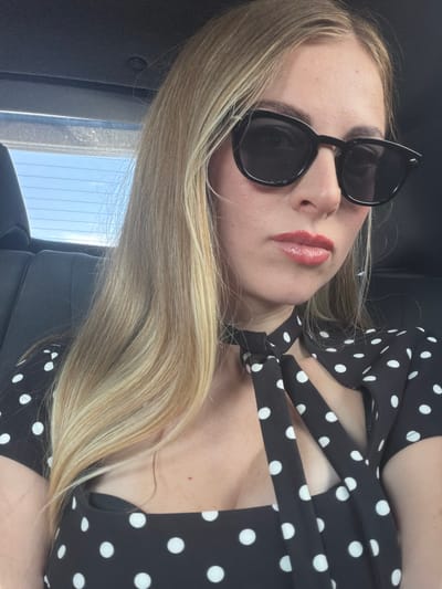 A woman with blonde hair and sunglasses is taking a selfie in the back seat of a car. She is wearing a black and white polka dot dress. The background shows the interior of a car, with the window behind her revealing a blue sky.
