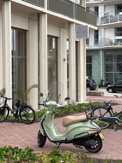 In Amsterdam, Netherlands, a light green Vespa scooter is parked on a brick walkway in front of a modern building with concrete pillars and large windows. A black bicycle is parked to the left of the scooter, and another black bicycle is partially visible to the right. In the background, a black car and a black motorcycle are parked on the street. A sign for "Rinki Kappers" is attached to the building above the scooter. Green plants are growing in front of the walkway.