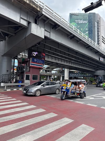 This image depicts a street scene in Bangkok, Thailand, likely during the daytime. In the foreground, a red and white striped pedestrian crossing is visible, with a gray sedan stopped on the left side of the frame and a blue and yellow tuk-tuk vehicle with three passengers in the center. The tuk-tuk appears to be in motion, navigating the intersection. Behind the vehicles, an elevated structure, possibly part of an expressway or bridge, dominates the upper portion of the image. A modern glass-fronted building with a green advertisement banner displaying Thai text and an image of elephants is visible on the right, suggesting a commercial or business district. To the left of the pedestrian crossing, a small, maroon booth with stairs leading up to it is situated under the elevated structure, with a Thai flag and a digital billboard displaying text and images above it. Traffic signals and other street infrastructure are also present in the background, indicating a busy urban environment. The sky is overcast, suggesting cloudy weather.