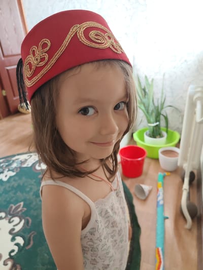 A young girl with brown hair is looking at the camera, smiling. She is wearing a red fez with gold embroidery and a white floral dress. The background is blurry, but it appears to be an indoor setting with a wooden floor and a rug. There are several objects in the background, including plants in pots, a red bucket, and a white radiator. The lighting suggests it is daytime. The overall mood is cheerful and playful. The girl is likely posing for the photo.