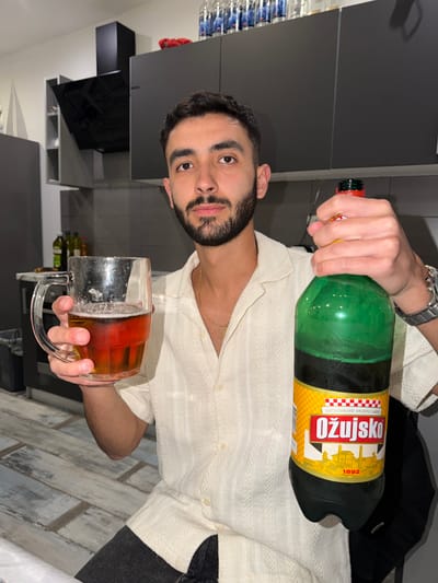A young man with dark hair and a beard, wearing a light-colored, textured collared shirt and a gold chain, looks directly at the camera. He wears a watch on his left wrist. The image is taken indoors, likely in a kitchen or a home setting in Grad Rijeka, Croatia, identifiable by the prominent brand of beer.
In his right hand, he holds a large, green plastic bottle of "OŽUJSKO" beer, a Croatian brand, with a red cap. The yellow and red label clearly displays "OŽUJSKO" in bold red letters, along with the text "OSVJEŽAVAJUĆI SVIJETLI LAGER" (Refreshing light lager) and the year "1892", accompanied by an illustration of buildings. His left hand grasps a large, clear glass mug, partially filled with an amber liquid and a layer of foam, indicative of beer.
The background features modern, dark grey cabinetry or shelves, suggesting a contemporary interior. To the far left, a stack of bottled water is visible. A dark fabric item, possibly clothing, rests behind the beer bottle. The floor below appears to be light-colored, with a distressed or wood-grain pattern. The man's expression is neutral, with a slight hint of a smile, suggesting a casual and relaxed moment, likely enjoying a beverage.