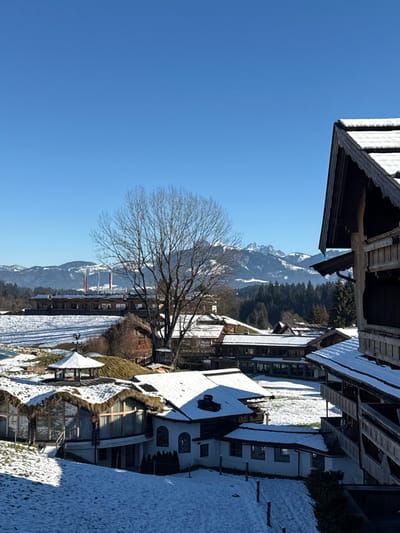 The image depicts a snow-covered alpine landscape in Going am Wilden Kaiser, Austria, under a clear, unclouded blue sky. In the immediate foreground to the right, a section of a multi-story traditional wooden building with a dark, snow-dusted gabled roof and ornate carved wooden balconies is visible. The midground features a cluster of buildings partially covered in snow. A prominent structure exhibits a curved facade with extensive glazing, a textured, possibly grass-covered or thatched roof, and is partially set into a snowy slope. Adjacent are white-walled buildings with dark, snow-covered roofs. A small, white hexagonal gazebo with a dark conical roof topped by a weathervane is situated in the lower-left midground. A large, bare deciduous tree stands centrally, its branches reaching towards the sky. The ground throughout the scene is covered in a layer of white snow, with some patches of brown vegetation visible. In the background, a large, elongated building with multiple snow-covered sections extends horizontally on a distant snowy incline. Further in the distance, a dense coniferous forest line transitions to rugged, snow-capped mountains of the Wilder Kaiser range. On the distant left horizon, behind the elongated building, three tall, red-and-white striped industrial chimneys rise prominently. The scene is illuminated by bright sunlight, casting distinct shadows. No individuals or animate subjects are present.