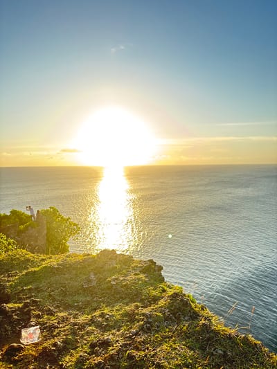 The image shows a landscape at Pecatu, Indonesia during sunset. The sun is setting over the ocean, creating a bright reflection on the water's surface. The sky is blue, fading to yellow near the horizon. The cliff is covered in green grass, a partially transparent plastic package lying on the grass. There is a person standing at the edge of the cliff, looking towards the ocean. The lighting suggests it is golden hour.
