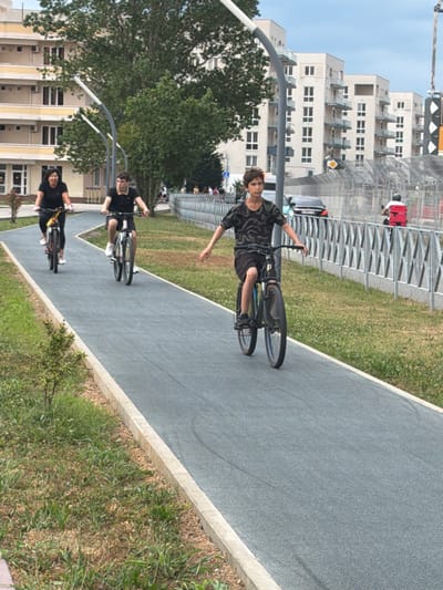 In Sirius, Russia, on a clear day, three individuals are cycling on a dedicated asphalt bike path. In the foreground, a young person with short brown hair, wearing a dark patterned t-shirt and black shorts, rides a dark bicycle with a blue frame. Their arms are outstretched, and they are looking forward. Behind them, slightly to the left, a person in a black t-shirt and dark pants rides a dark bicycle. To the left of that, a woman with dark hair, wearing a black t-shirt and white shoes, rides a yellow and black bicycle. The bike path is bordered by green grass and a concrete curb. In the background, there are modern apartment buildings with multiple balconies and windows. A metal fence runs along the right side of the bike path, and beyond that, there is traffic including a black car and a scooter with a red delivery box. The sky is partly cloudy.