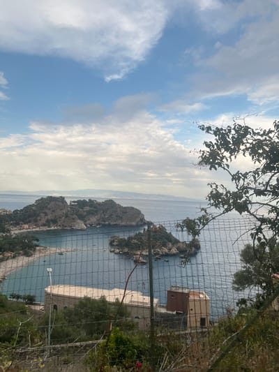 This is a landscape photograph taken from an elevated vantage point, overlooking a coastal scene in Taormina, Italy. The image captures a bay with a small island, known as Isola Bella, at its center. The sea is a deep blue, with calm waters. Isola Bella is a rocky outcrop covered in lush green vegetation, with a narrow causeway connecting it to the mainland.

In the foreground, a wire fence partially obstructs the view, with green foliage and dry branches visible through the mesh. Beyond the fence, on the mainland, a section of a building with a light-colored roof and light brown and beige walls is visible. The building appears to be of stone construction. Further inland, a sandy beach is dotted with people, and several small boats are anchored in the bay.

The backdrop consists of rolling hills covered in trees, and a distant coastline stretches across the horizon. The sky is a mix of blue and white clouds, suggesting it is either morning or late afternoon. The overall impression is of a serene coastal landscape with a touch of natural and man-made elements. The specific location is likely the viewpoint from which Isola Bella and the Taormina coastline are famously observed.