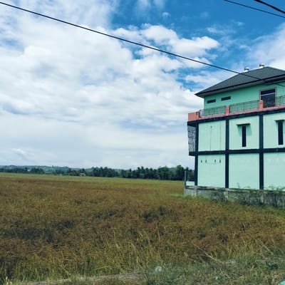 This image captures a vibrant daytime scene in Cotbada, Indonesia, featuring a distinctive building alongside a vast, uncultivated field.

The primary subject on the right side of the frame is a multi-story building painted in a prominent mint green color. It features strong architectural lines with black framing around its windows and structural elements, giving it a somewhat industrial or modern appearance. The roof is dark green, and a balcony on the upper floor is accented with reddish-orange and has a black railing. The top of the roof shows two circular vents. A section of the building on the left side features a caged or barred structure. The building is bordered by a low concrete wall at its base.

To the left and extending into the foreground and midground is a wide expanse of field, covered in tall, dry, brownish-green grass and weeds, suggesting either uncultivated land or a dry season. In the distant background, a line of lush green trees, including what appear to be palm trees, separates the field from gently rolling hills under a slightly hazy sky. A small house with a red roof can be faintly seen nestled among the distant trees.

The sky is a bright, clear blue with scattered white, fluffy clouds, indicating fair weather. Two dark utility lines stretch diagonally across the upper portion of the sky, adding a modern element to the natural and architectural landscape. No human activity or visible text is present in the image. The scene is static, showcasing the intersection of built environment and natural landscape under a bright, open sky.