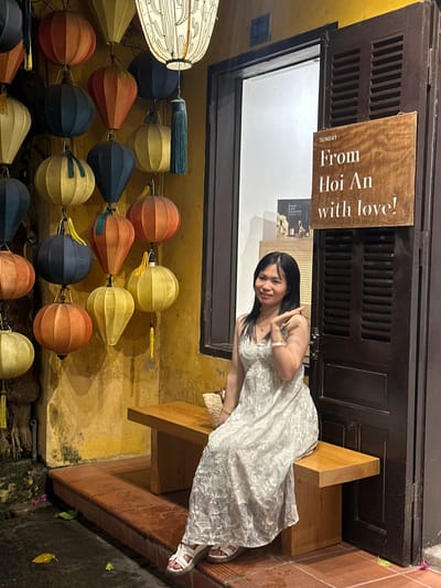 In a nighttime scene, a young woman in a white lace dress sits on a wooden bench outside a shop. She is smiling and making a peace sign with her fingers. Behind her, a dark wooden door with louvers is visible, with a brown sign hanging from it that reads "SUNDAY From Hoi An with love!". Below the door, a row of colorful, round paper lanterns hangs, illuminated by an unseen light source, creating a warm and inviting ambiance. The lanterns are a mix of yellow, orange, blue, and red, with some featuring decorative tassels. The wall behind the lanterns is a textured yellow, suggesting an outdoor setting in a location with traditional architecture. The ground appears to be dark and wet, possibly indicating recent rain. The overall mood is cheerful and relaxed, capturing a moment of enjoyment during a trip to Hội An.