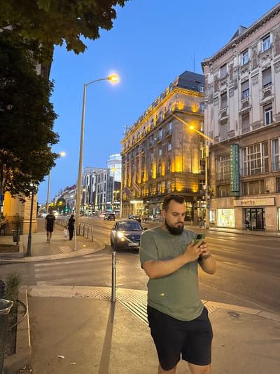 A man, with a beard and short brown hair, stands on a sidewalk in a city at dusk. He is wearing a green t-shirt and dark blue shorts and is looking at his phone, which he holds in his right hand. The street is illuminated by streetlights and the lights from buildings. Several cars are visible on the street, and there are pedestrians walking on the sidewalk. The buildings appear to be old, with ornate architecture. The sky is a deep blue, indicating that it is twilight. The overall mood is peaceful and urban.
The image appears to be taken in Budapest, Hungary, given the context provided. The name "ASTORIA" is visible on a sign on one of the buildings, which is likely referring to the Astoria intersection or a building in that area of Budapest. There are other signs in Hungarian, although they are not clearly legible. The time of day is dusk, with streetlights and building lights beginning to illuminate the scene. The weather appears to be clear.