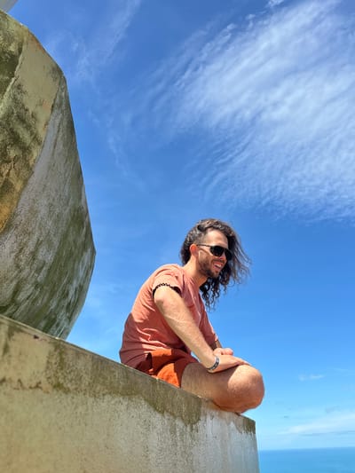 A man with long, curly brown hair and sunglasses is sitting cross-legged on a concrete ledge, looking to his right and smiling. He is wearing an orange t-shirt and orange shorts. Behind him, the sky is a bright blue with wispy white clouds. To his left, a curved concrete structure, possibly part of a building or monument, is visible, showing signs of age and weathering with patches of green moss. The background shows a clear view of the blue ocean. The setting appears to be an elevated outdoor location, offering a panoramic view of the sea. This location could be a viewpoint or a rooftop on Ko Samui, Thailand, given the tropical weather and ocean vista.