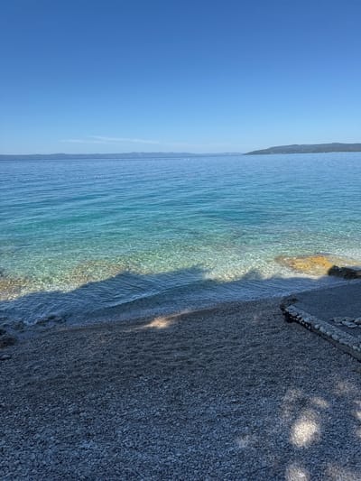 The image shows a serene view of the Adriatic Sea in Makarska, Croatia. A clear, turquoise sea stretches out towards a distant mountainous coastline, under a bright blue sky. The water is calm, with gentle waves lapping at a pebble beach in the foreground. The beach is composed of small, smooth pebbles, with a paved path leading away from the water's edge, bordered by a low stone wall. The water is so transparent that the sandy and pebbly seabed is visible. A line of buoys, marking a swimming area, stretches across the middle distance of the sea. The scene evokes a sense of tranquility and natural beauty.