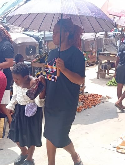The image depicts a busy outdoor market scene in Ura Nta Umuarandu, Nigeria. In the foreground, a woman and a child walk from left to right. The woman, positioned slightly ahead and to the right, has medium-dark skin and reddish-brown braided hair styled in a ponytail. She wears a black short-sleeved dress featuring a colorful "GUCCI" logo on the chest and carries a light purple/grey polka-dotted umbrella with a wooden handle in her right hand, providing shade. She wears dark sandals.
To her left and slightly behind is a child, a girl with short dark curly hair and medium-dark skin. She wears a white long-sleeved blouse with ruffled cuffs, a black skirt, and grey Crocs-style shoes. The child holds a small, translucent dark-colored purse with a gold chain strap in her right hand and has a small, round, purple patterned bag with a chain strap slung across her body.
The background reveals a bustling market environment. To the right, a pile of small orange-red fruits, possibly garden eggs, is displayed on a light-colored cloth directly on the ground. Behind this, a wooden market stall supports green bananas. Another person, partially visible, walks away in the far right. In the mid-ground and left, several three-wheeled auto-rickshaws (Keke Napep) are parked or moving, some bearing license-like numbers such as "AD528" and text like "ASTORY". Other market stalls, shaded areas from canopies, and blurred figures of additional people are visible, indicating a crowded setting under bright daylight. The ground appears to be a mix of concrete and dirt.