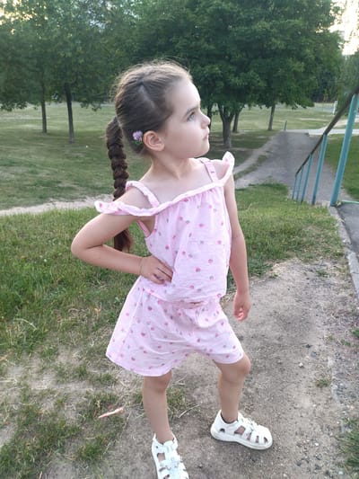 A young girl with brown hair in a braid, wearing a pink outfit with a strawberry pattern, stands on a dirt path. She is looking to her right, with her left hand on her hip. Her hair has a small purple flower tucked into it. In the background, there are trees and a grassy area. A blue railing and stairs are visible to the right. The location is a park in Hrodna, Belarus.